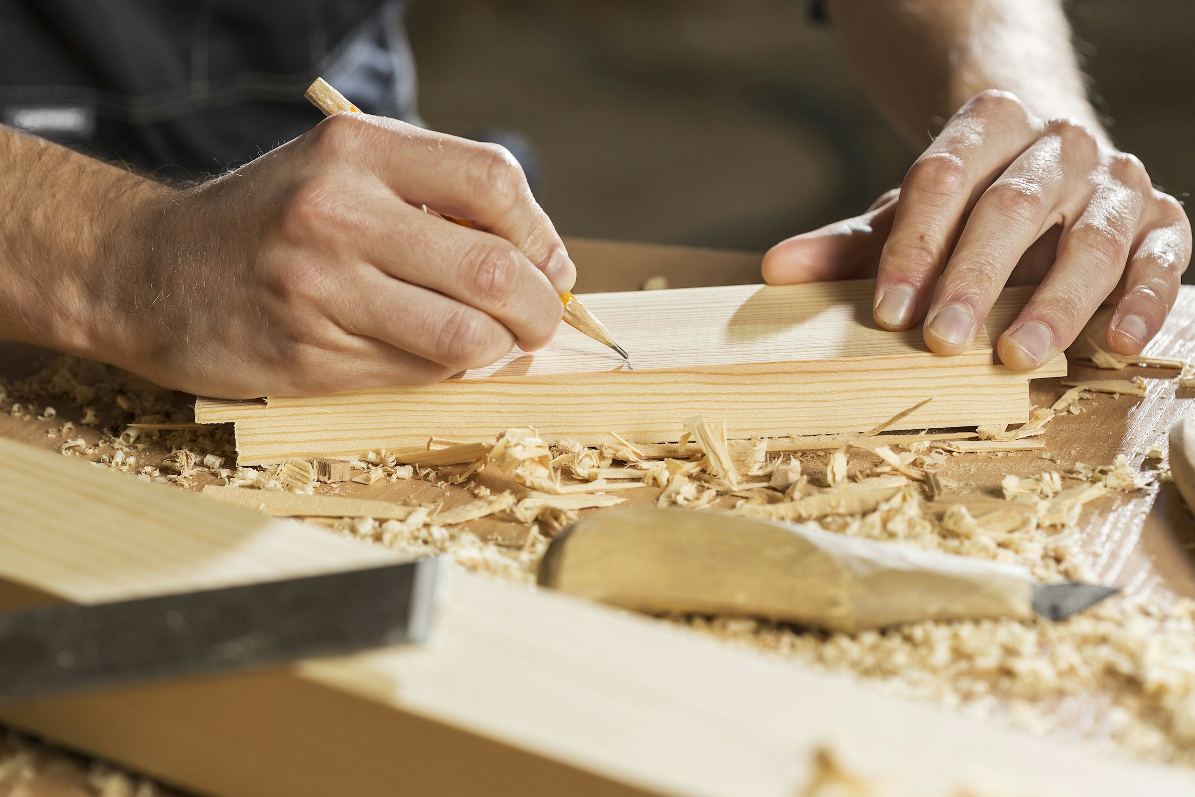 Handwerker markiert ein Werkstück aus Massivholz mit dem Bleistift, umgeben von Hobelspänen